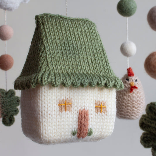 Hand-knitted cream house with a scalloped sage green roof and embroidered window details.
