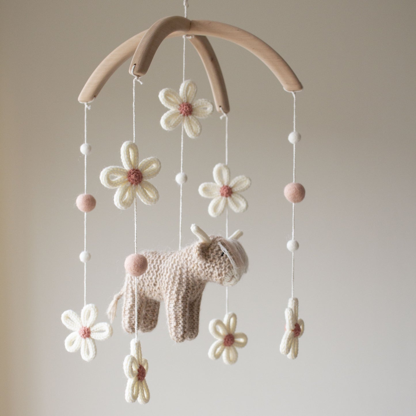 Hand-knitted highland cow baby mobile with ivory daisies and dusty rose wool balls on a wooden hanger.
