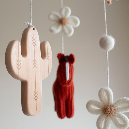 Close-up of two hand-knitted horses in rich brick red wool for a western-themed nursery. Detailed shot of a carved wooden cactus and tee-pee charms on an artisan baby mobile.