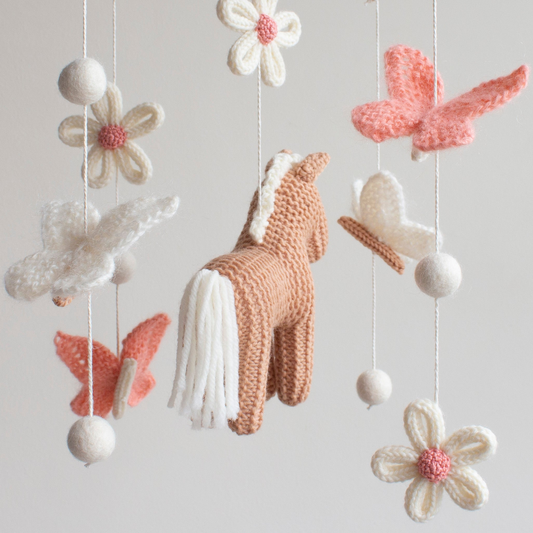 Knitted light brown pony with cream mane and tail, surrounded by fluttering daisies and and butterflies hanging on a artisan baby mobile