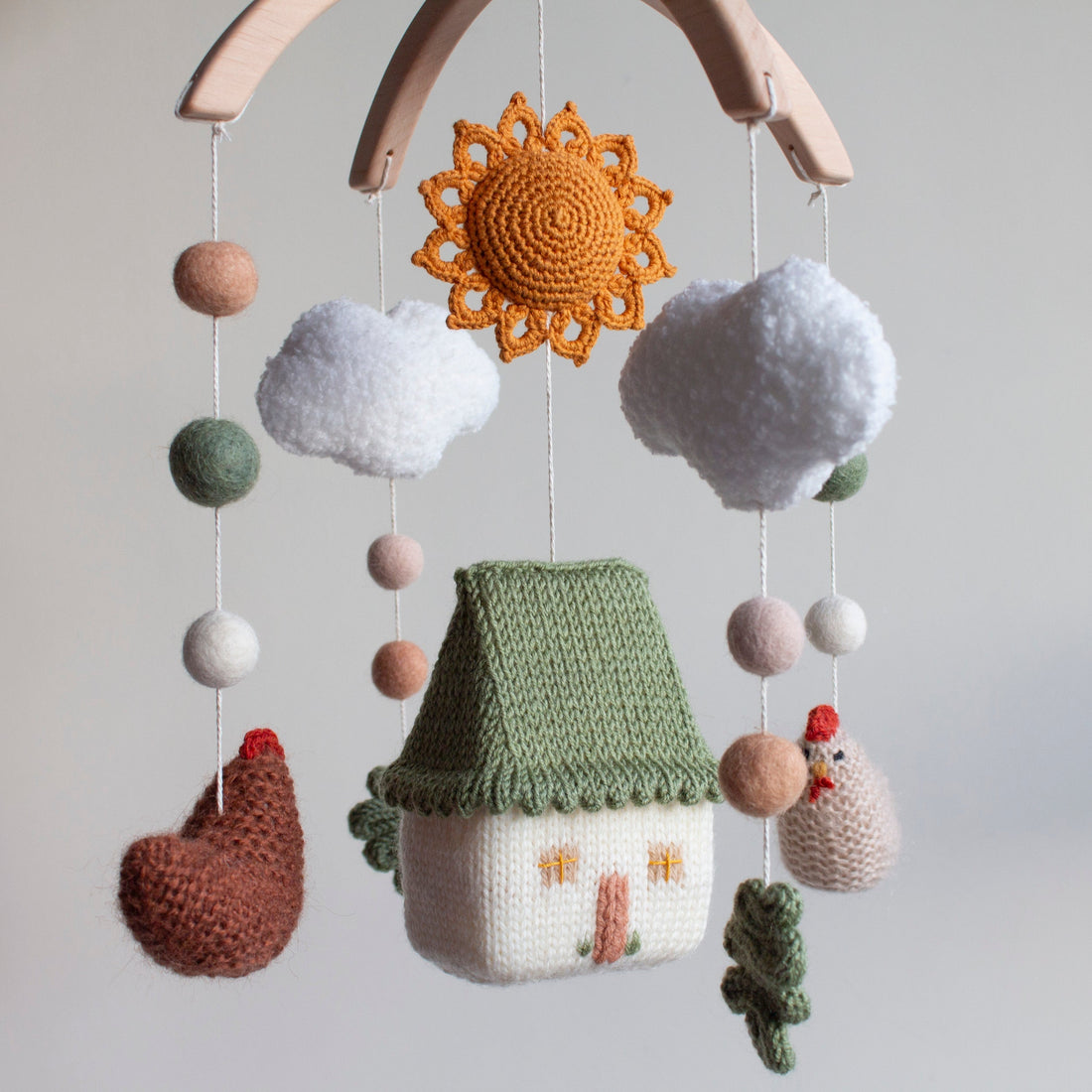 Hand-knitted farmhouse baby mobile featuring a cream cottage with a scalloped sage green roof, cinnamon brown and beige chickens, a vibrant burnt orange sun, and fluffy white boucle clouds suspended from a natural wood arch.