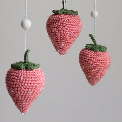 Close-up of a hand-crocheted coral strawberry amigurumi with green leaves for a fruit-themed nursery. Macro view of 100% natural wool yarn stitches on a handmade artisan strawberry mobile.