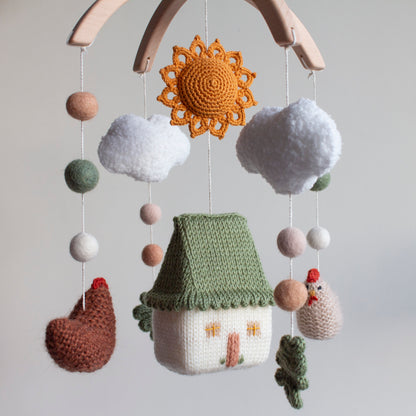 Hand-knitted farmhouse baby mobile with a green-roofed cottage, cinnamon brown chickens, and a burnt orange sun.