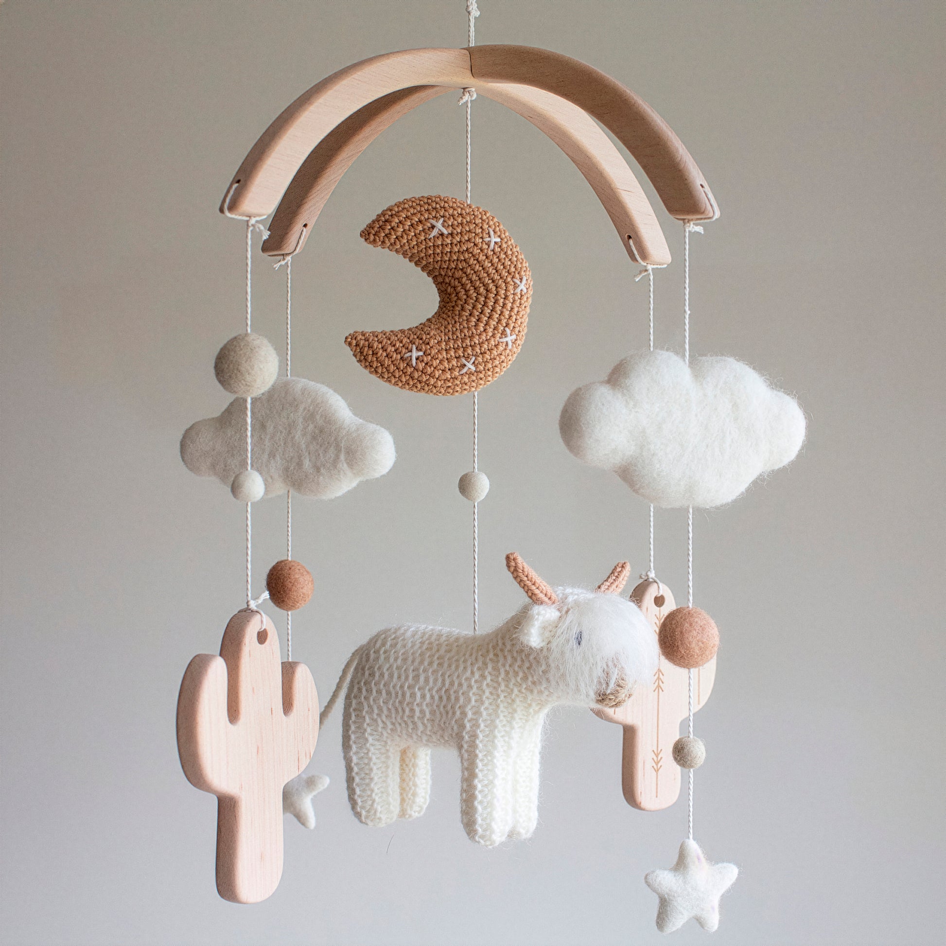 Side view of a whimsical ruralism baby mobile featuring a shaggy white cow and untreated solid alder wood cactuses. Hand-stitched with high-quality wool yarn and cotton yarn, this mobile offers a palette of soft whites and earthy browns. The untreated solid wood mobile frame provides clean lines for a modern nursery. A perfect choice for those seeking a high-quality heirloom gift, its timeless design ensures it is suitable for different baby gifts and modern nursery themes.