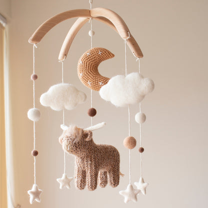 Hand-knitted highland cow baby mobile with crochet moon and white clouds on a wooden arch. High-contrast white stars and dark highland cow elements designed to support infant visual stimulation.