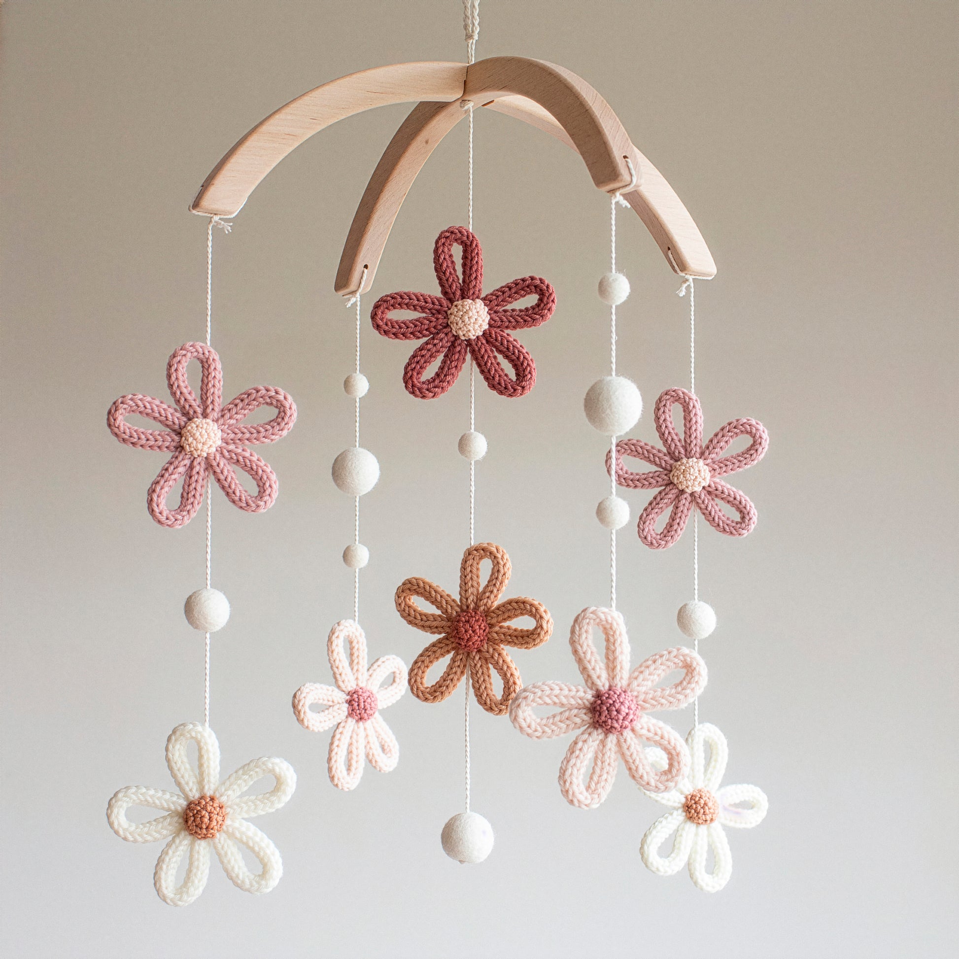 A wide shot of a handmade crochet flower mobile with eight blooming daisies in shades of terracotta, dusty rose, and cream. Each flower is meticulously hand-stitched using wool yarn OEKO-TEX Standard 100 safe for nurseries. The untreated solid wood hanger adds a rustic touch to the timeless design. This versatile piece is suitable for different baby gifts, including gender-neutral nursery decor. Its durable construction makes it a cherished heirloom gift that lasts through the early years.
