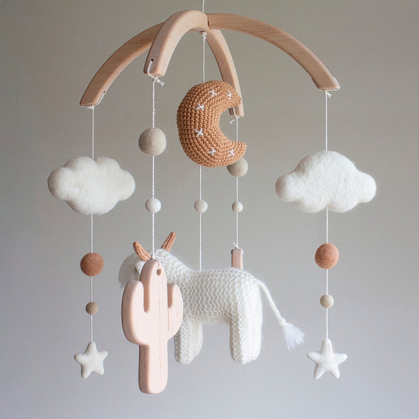 A perspective shot of the "Whimsical Ruralism" mobile, highlighting the textural juxtaposition between the shaggy cow and smooth untreated solid wood cactuses. Hand-stitched with wool yarn and cotton yarn, the elements hang from a sturdy untreated solid wood mobile frame. This timeless design is crafted in the UK to be a cherished heirloom gift. Its natural materials and neutral palette make it suitable for different baby gifts, fitting seamlessly into various nursery decors.