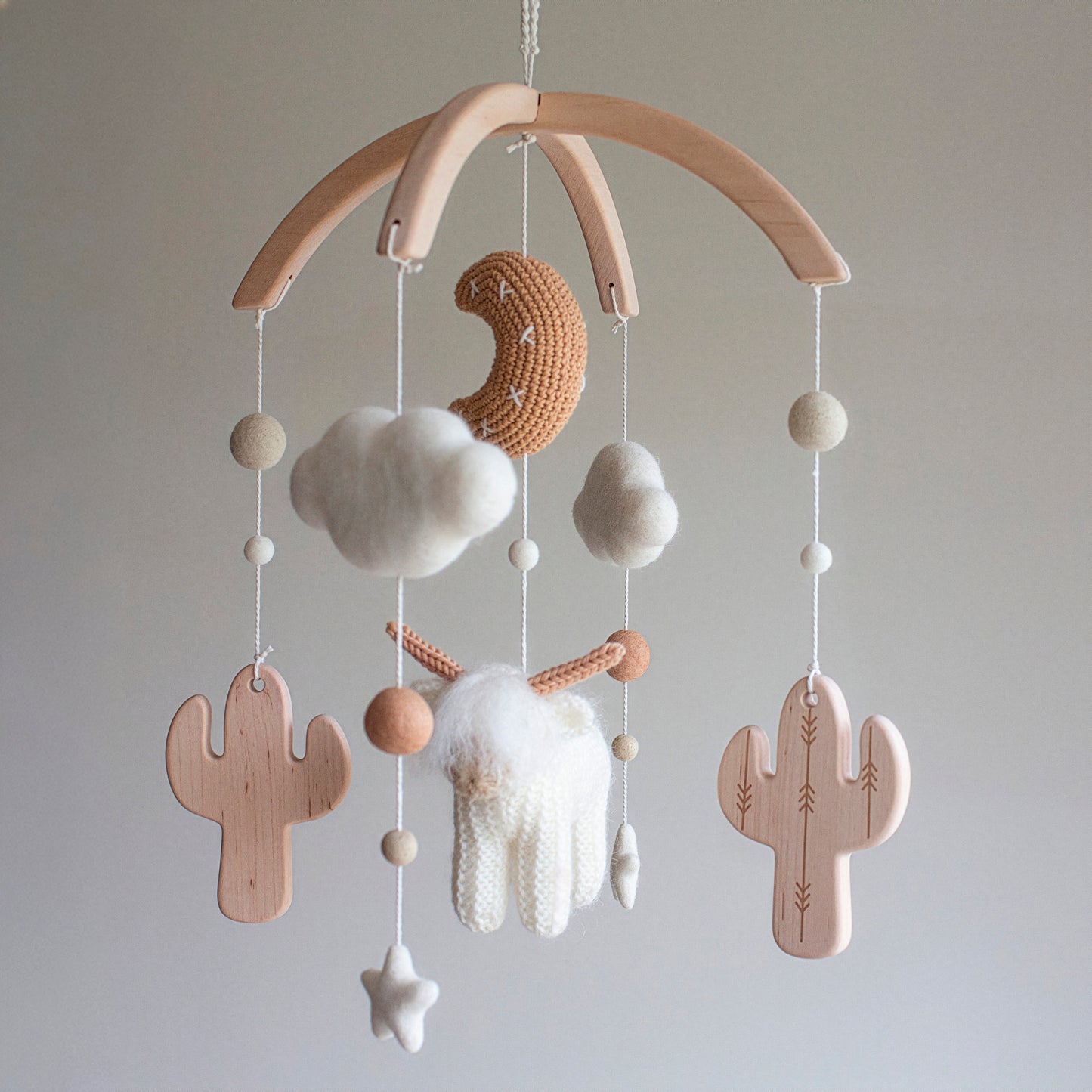 A beautiful White Longhorn Cow & Wooden Cactus Mobile featuring a tan moon with meticulous white stitches. The elements are handmade with wool yarn OEKO-TEX Standard 100 safe for baby environments and suspended from an untreated solid wood mobile frame. Including untreated solid wood cactuses and felt clouds, this mobile represents a timeless design. It serves as a meaningful heirloom gift and is suitable for different baby gifts for those who value artisan heritage and quality.