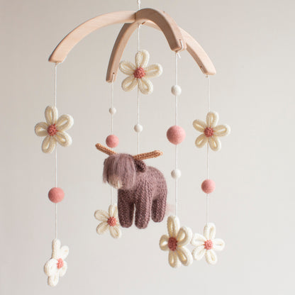 Hand-knitted highland cow baby mobile with ivory daisies and dusty rose wool balls on a wooden hanger