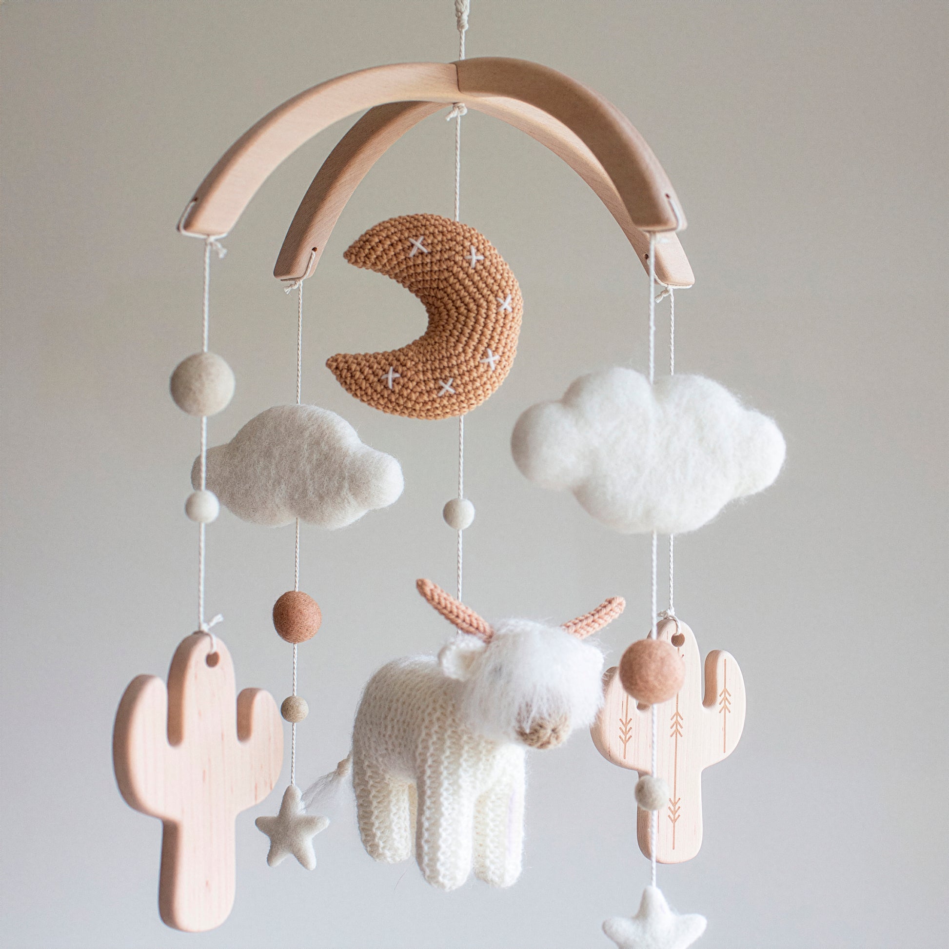 A White Longhorn Cow & Wooden Cactus baby Mobile capturing the minimalistic Scandi aesthetic. It features a shaggy, fluffy hand-knitted cow and a tan moon made of premium wool yarn OEKO-TEX Certified safe for newborns. Suspended from an untreated solid wood mobile frame, the design includes alder wood cactuses and soft wool felt clouds. This timeless design is an ideal heirloom gift, serving as a one-of-a-kind treasure suitable for different baby gifts.