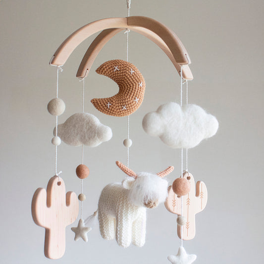 A White Longhorn Cow & Wooden Cactus baby Mobile capturing the minimalistic Scandi aesthetic. It features a shaggy, fluffy hand-knitted cow and a tan moon made of premium wool yarn OEKO-TEX Certified safe for newborns. Suspended from an untreated solid wood mobile frame, the design includes alder wood cactuses and soft wool felt clouds. This timeless design is an ideal heirloom gift, serving as a one-of-a-kind treasure suitable for different baby gifts.