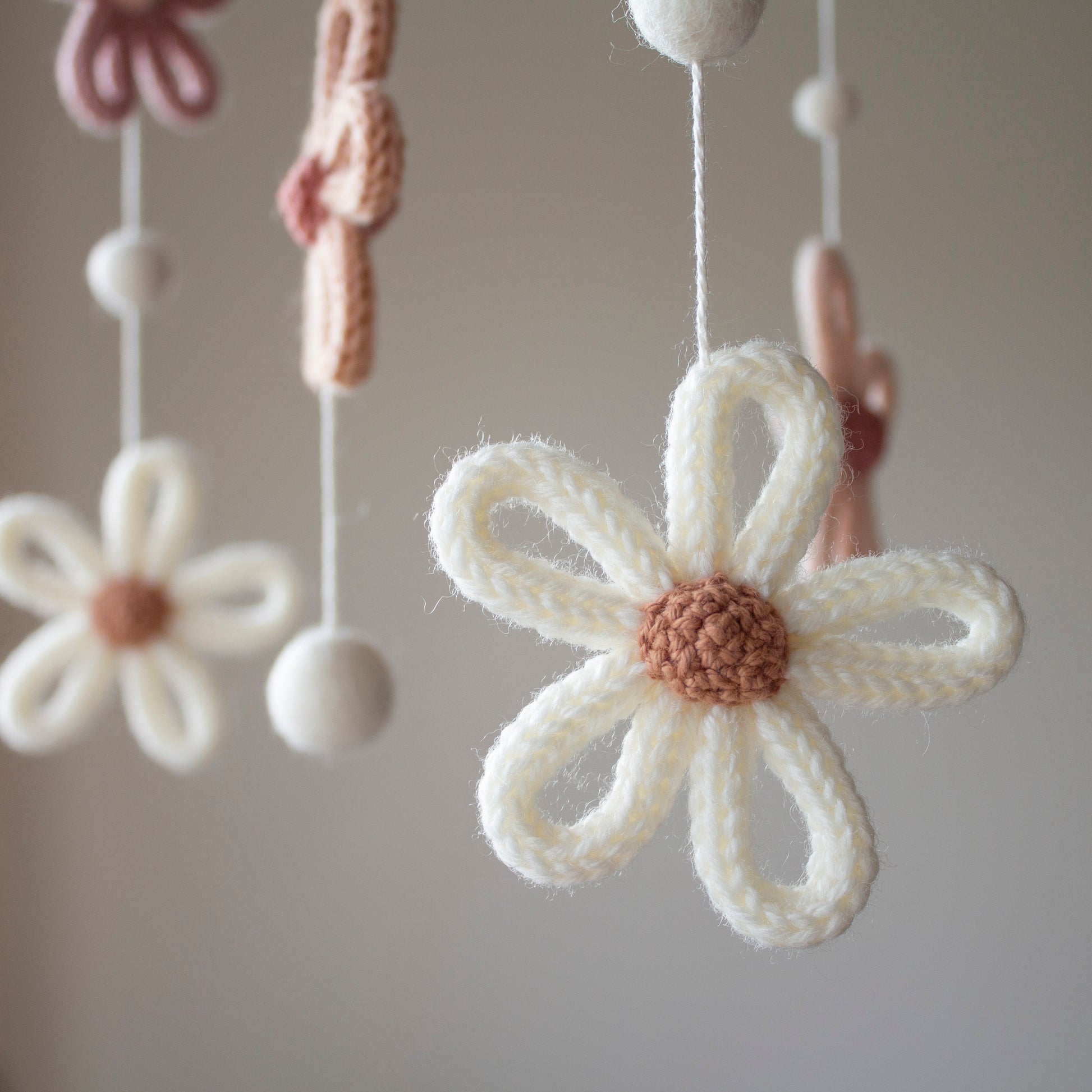 A detailed close-up of a cream-colored knitted flower with a brown center, hanging elegantly on a mobile string. The use of wool yarn OEKO-TEX Standard 100 safe for nurseries ensures a healthy environment for babies. This mobile features untreated solid wood, lending a natural feel to its timeless design. Whether you are looking for a special heirloom gift or are shopping for suitable for different baby gifts, this handmade piece from Lanacrocheting.com offers style and safety.
