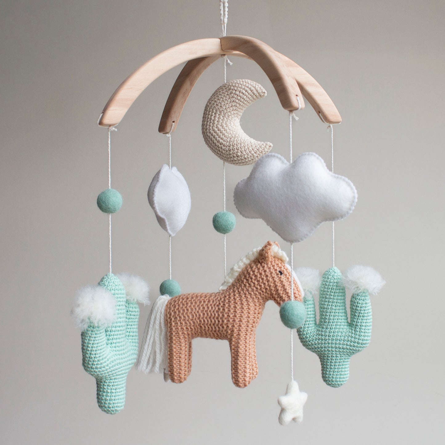 Artisan Western baby mobile featuring a tan hand-knitted horse, two mint green crochet cacti with white wool pom-poms, and a beige moon. Suspended from a sustainable wooden arch with ivory wool stars and clouds, this modern gender-neutral nursery decor is handcrafted in the UK from non-toxic materials. A prestigious heirloom-quality baby shower gift perfect for style-conscious mums-to-be looking for unique, ethical, and minimalist Scandi-inspired nursery accessories.