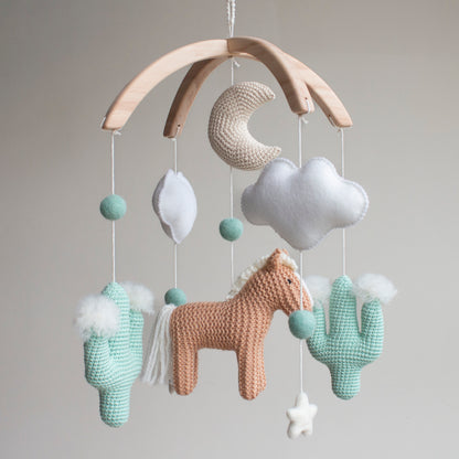 Artisan Western baby mobile featuring a tan hand-knitted horse, two mint green crochet cacti with white wool pom-poms, and a beige moon. Suspended from a sustainable wooden arch with ivory wool stars and clouds, this modern gender-neutral nursery decor is handcrafted in the UK from non-toxic materials. A prestigious heirloom-quality baby shower gift perfect for style-conscious mums-to-be looking for unique, ethical, and minimalist Scandi-inspired nursery accessories.