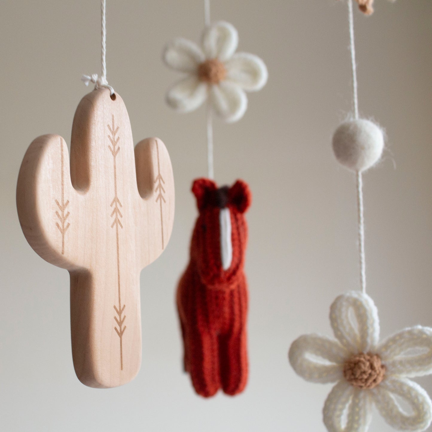 Close-up of two hand-knitted horses in rich brick red wool for a western-themed nursery. Detailed shot of a carved wooden cactus and tee-pee charms on an artisan baby mobile.