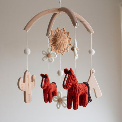 Hand-knitted boho cowgirl baby mobile with brick red horses and wooden cactus on a wooden hanger.