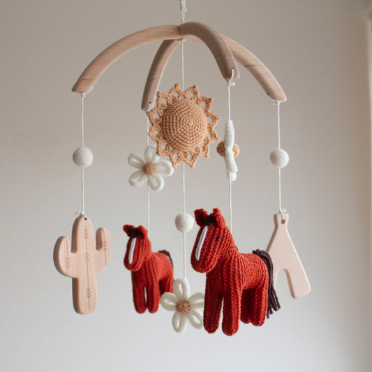 Hand-knitted boho cowgirl baby mobile with brick red horses and wooden cactus on a wooden hanger.