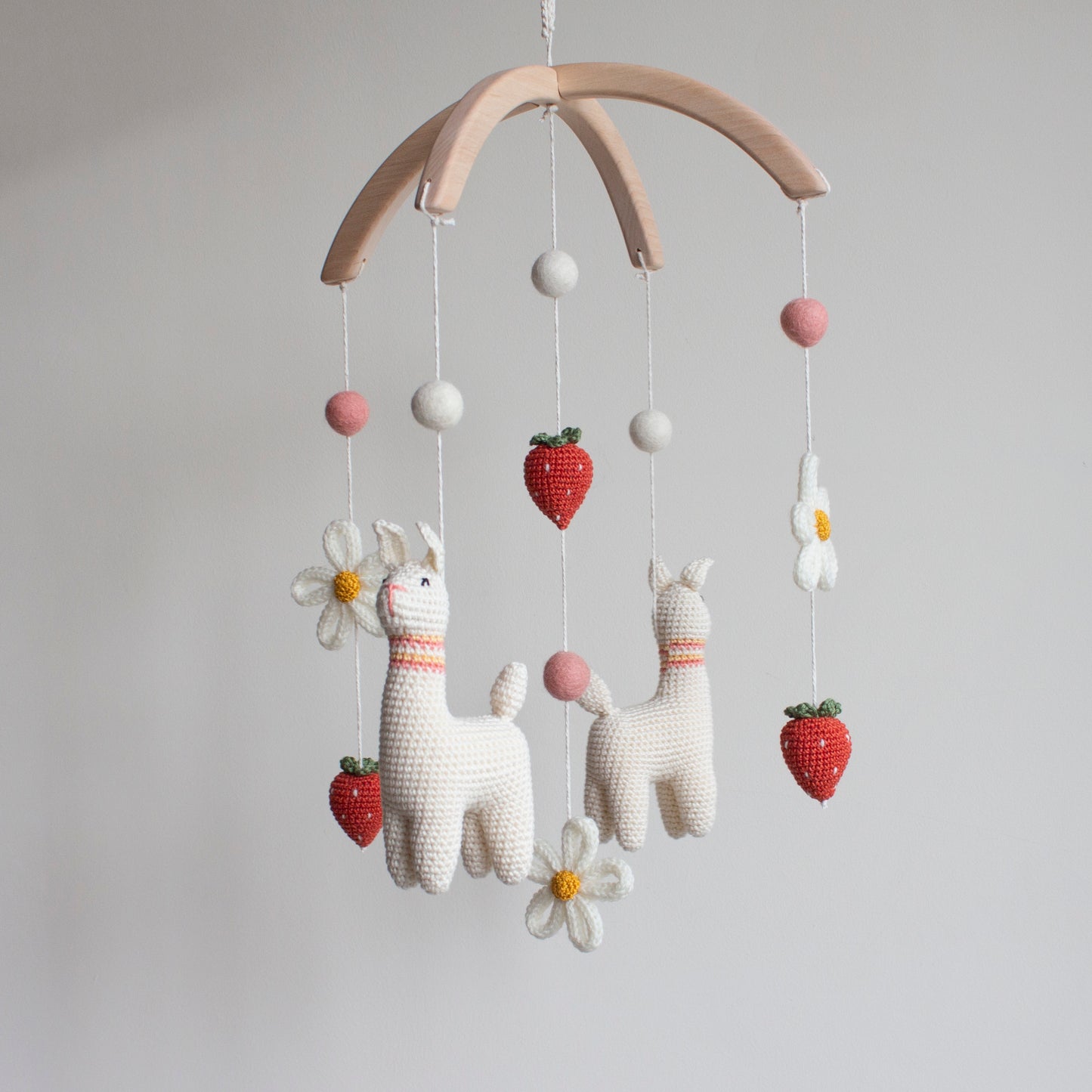 Baby mobile with white llamas, flowers, and strawberries hanging on a wooden arch. Unique boho nursery decor.
