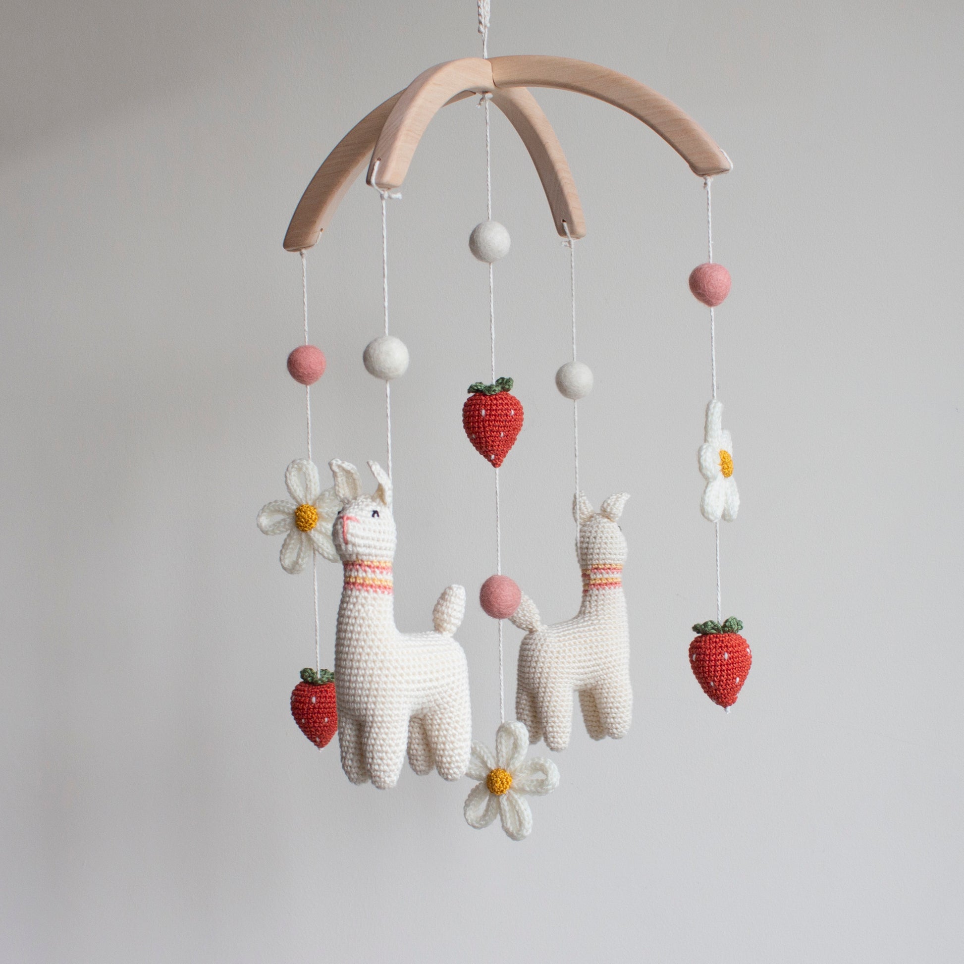 Baby mobile with white llamas, flowers, and strawberries hanging on a wooden arch. Unique boho nursery decor.