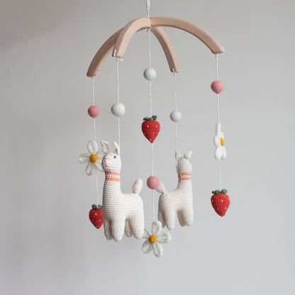 Baby mobile with white llamas, flowers, and strawberries hanging on a wooden arch. Unique boho nursery decor.