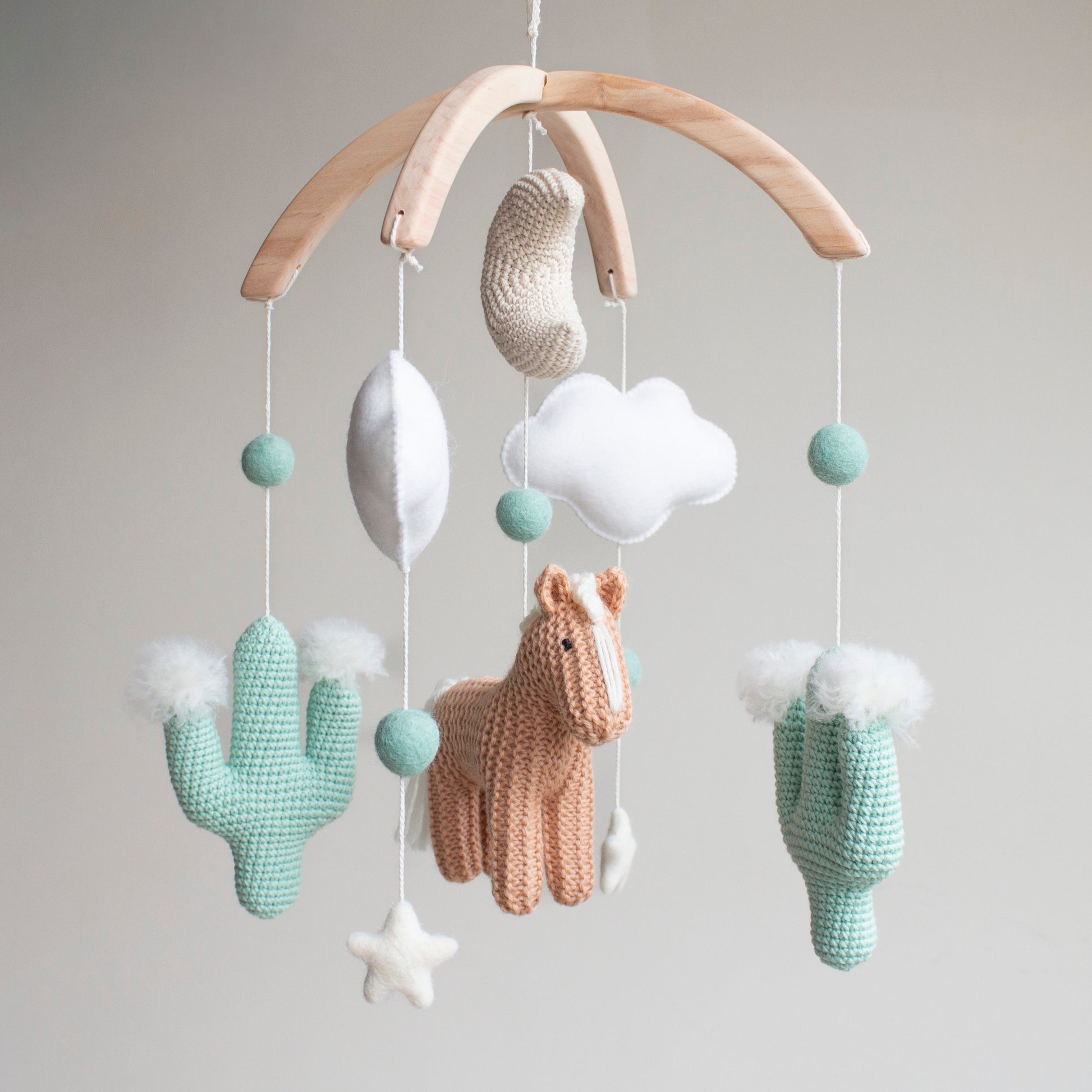 Artisan Western baby mobile featuring a tan hand-knitted horse, two mint green crochet cacti with white wool pom-poms, and a beige moon. Suspended from a sustainable wooden arch with ivory wool stars and clouds, this modern gender-neutral nursery decor is handcrafted in the UK from non-toxic materials. A prestigious heirloom-quality baby shower gift perfect for style-conscious mums-to-be looking for unique, ethical, and minimalist Scandi-inspired nursery accessories.