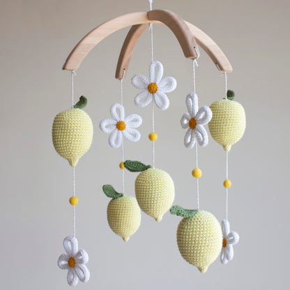 A handmade baby mobile featuring crocheted yellow lemons with green stems, white daisy flowers with yellow centers, and small yellow beads, suspended from a light wood arch against a neutral beige background.