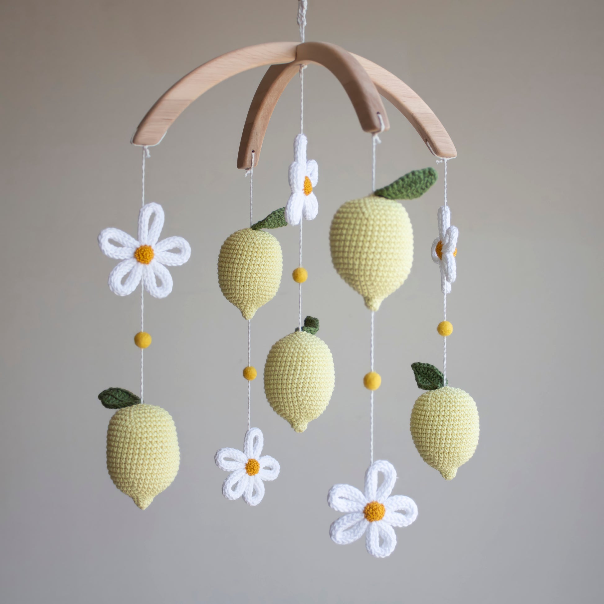 A handmade crochet baby mobile featuring yellow lemons with green leaves, white daisies with yellow centers, and small yellow beads, suspended from a curved natural wood arch against a neutral gray background.