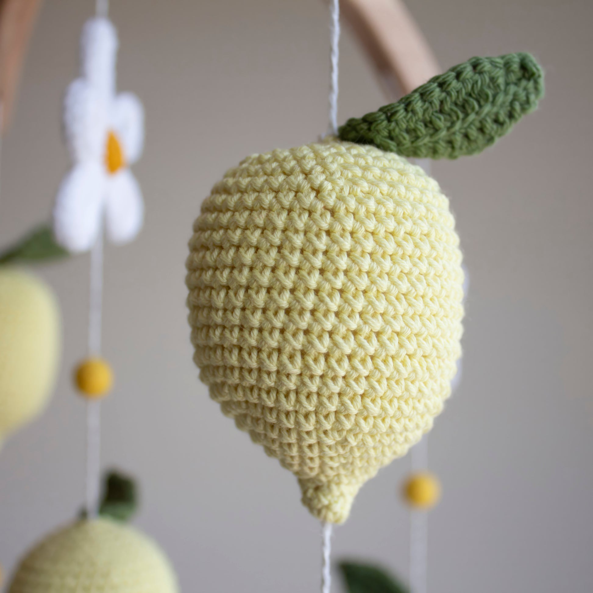 A close-up of a crocheted lemon hanging ornament in pale yellow with a green leaf, suspended against a softly blurred neutral background.