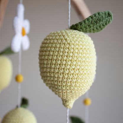 A close-up of a crocheted lemon hanging ornament in pale yellow with a green leaf, suspended against a softly blurred neutral background.