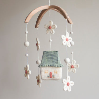 Hand-knitted Daisy Cottage baby mobile with sage green roof, white daisies, and blush pink accents on a wooden arch