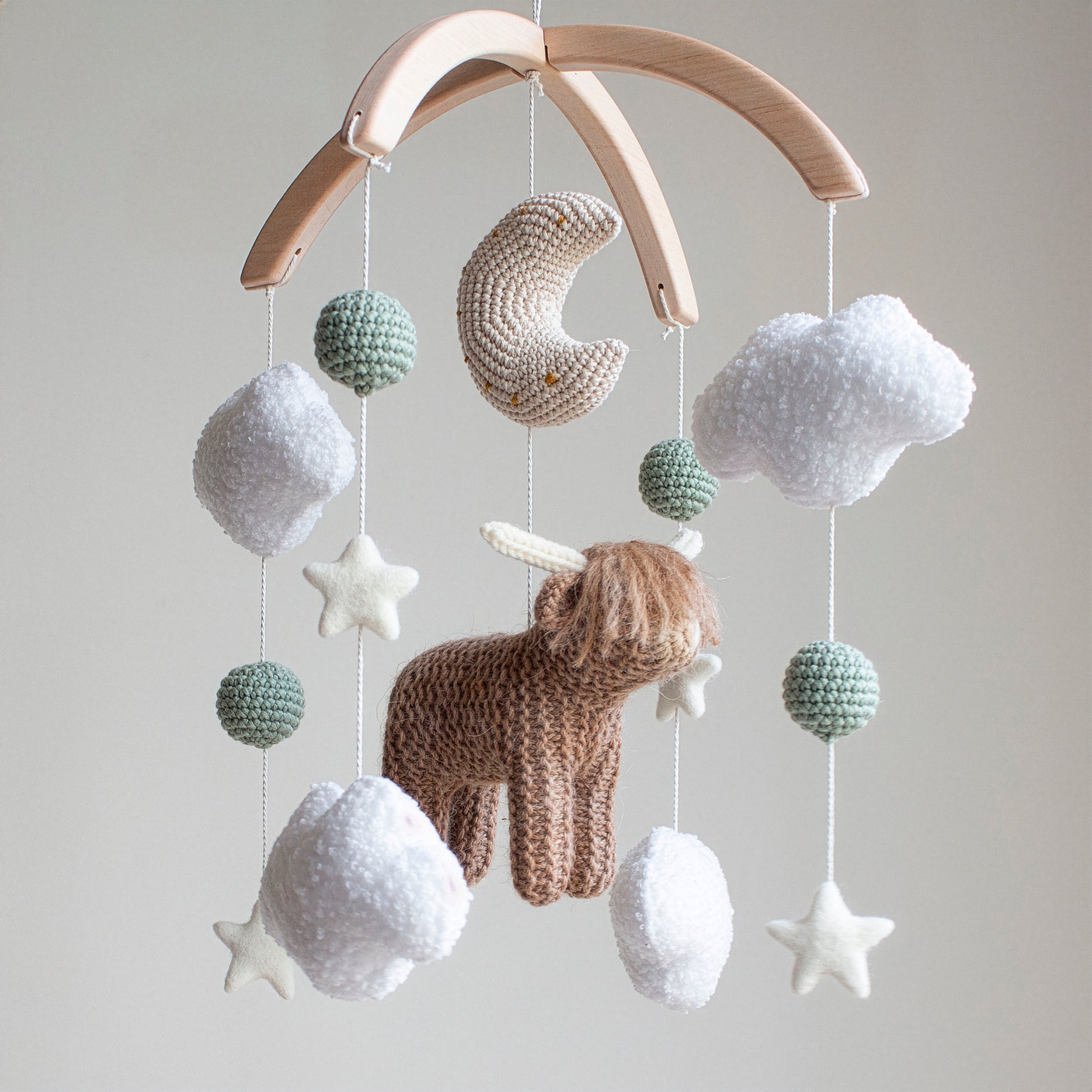 Hand-knitted highland cow baby mobile with white boucle clouds and sage green accents on a wooden arch.