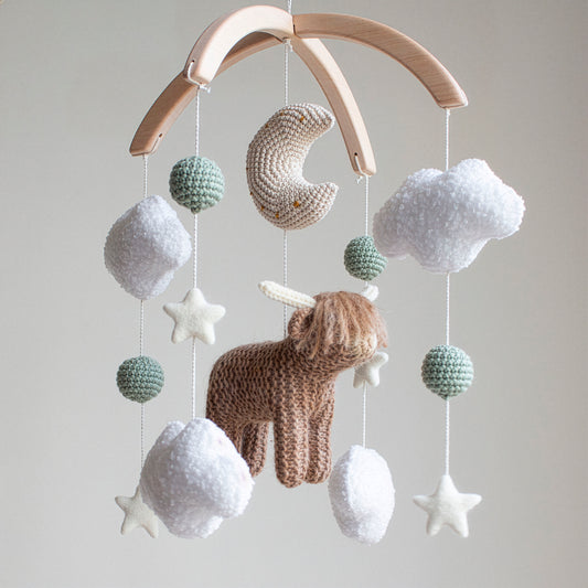 Hand-knitted highland cow baby mobile with white boucle clouds and sage green accents on a wooden arch.