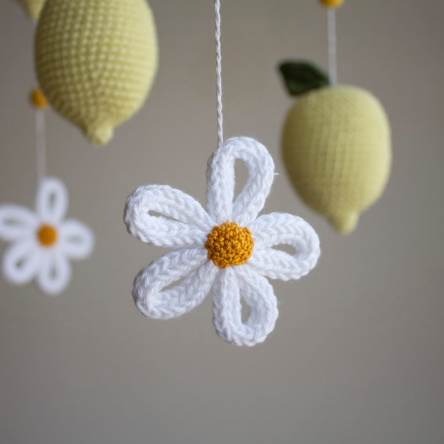 A close-up of a crocheted white daisy flower with a yellow center hanging from a white string, with two pale yellow crocheted lemons in the background against a soft gray wall.