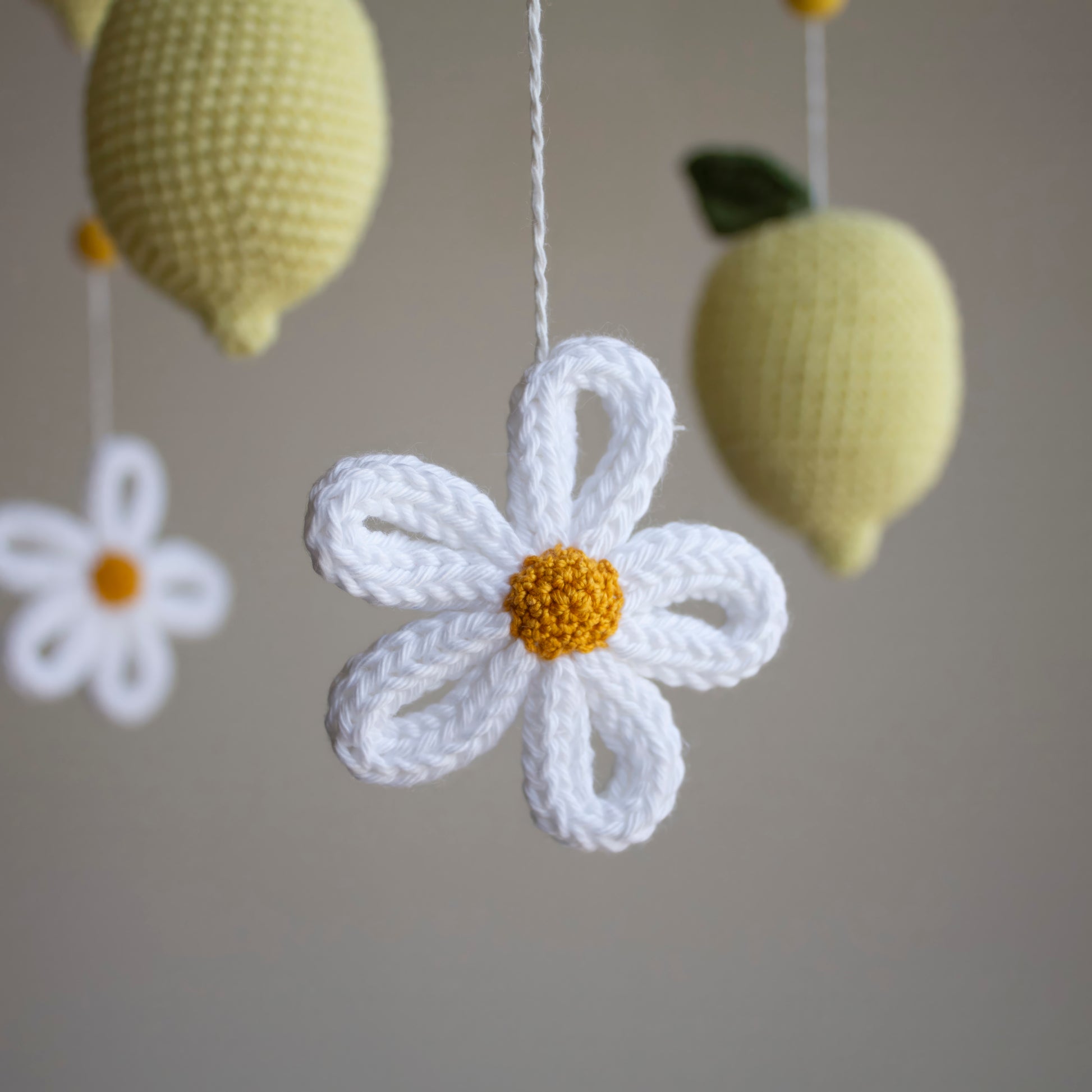 A close-up of a crocheted white daisy flower with a yellow center hanging from a white string, with two pale yellow crocheted lemons in the background against a soft gray wall.