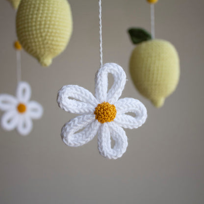 A close-up of a crocheted white daisy flower with a yellow center hanging from a white string, with two pale yellow crocheted lemons in the background against a soft gray wall.