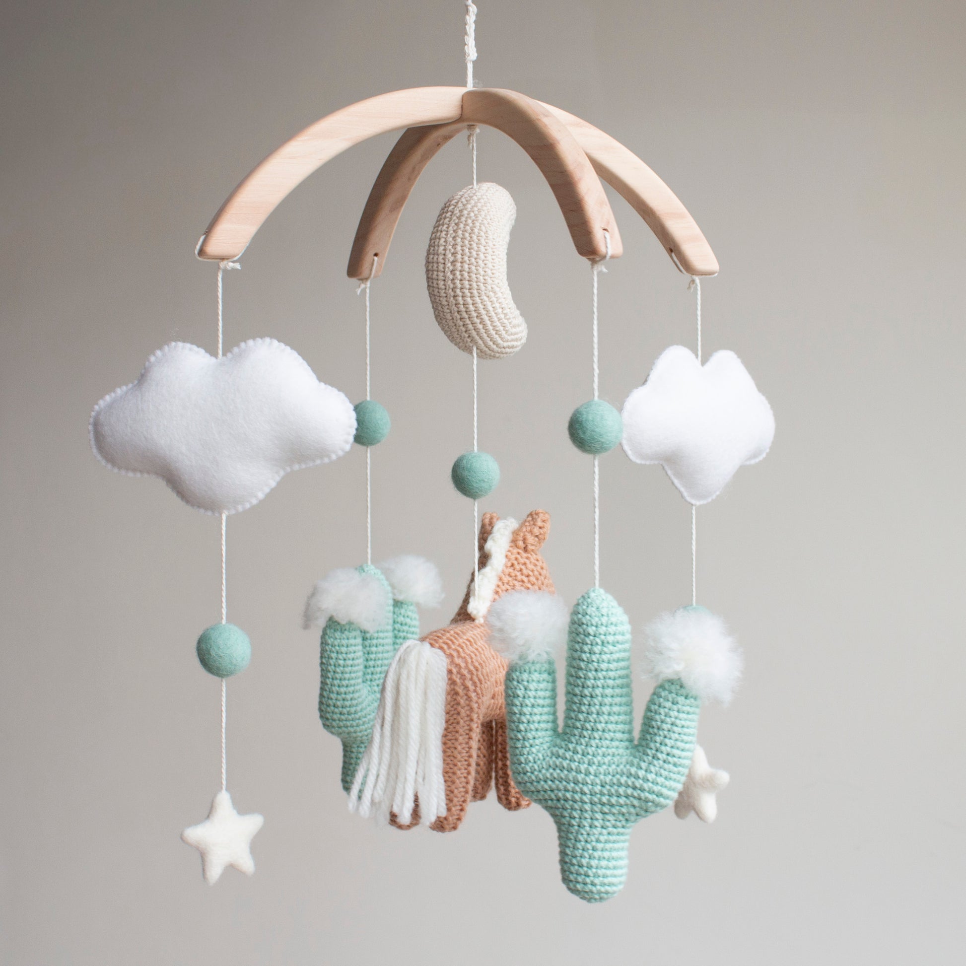 Handmade horse and cactus baby mobile in a muted mint, tan, and soft white palette. Every element is meticulously crafted using traditional "Slow Craft" techniques to ensure premium quality and durability. This nature-inspired accessory is a photogenic statement piece for a minimalist nursery, offering material transparency and safety for the baby's space. An ideal addition to UK baby registries, providing a high-end alternative to mass-market solutions for the discerning family