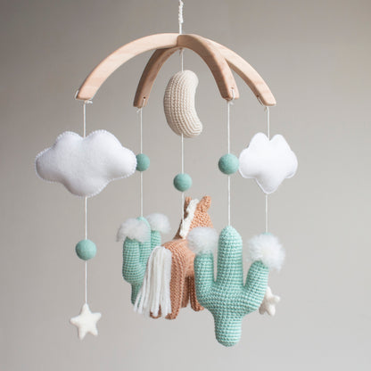 Handmade horse and cactus baby mobile in a muted mint, tan, and soft white palette. Every element is meticulously crafted using traditional "Slow Craft" techniques to ensure premium quality and durability. This nature-inspired accessory is a photogenic statement piece for a minimalist nursery, offering material transparency and safety for the baby's space. An ideal addition to UK baby registries, providing a high-end alternative to mass-market solutions for the discerning family