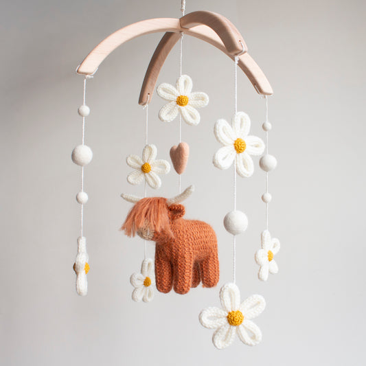 Nature-inspired rust-colored highland cow baby mobile with ivory knitted daisies and white wool clouds. This artisan nursery decoration is meticulously handcrafted in our Northern Ireland studio using certified organic wool. The earthy tones and botanical floral design make it a photogenic centerpiece for minimalist nurseries. A unique, vetted heirloom present that stands out from mass-market products for the discerning, high-net-worth UK consumer.