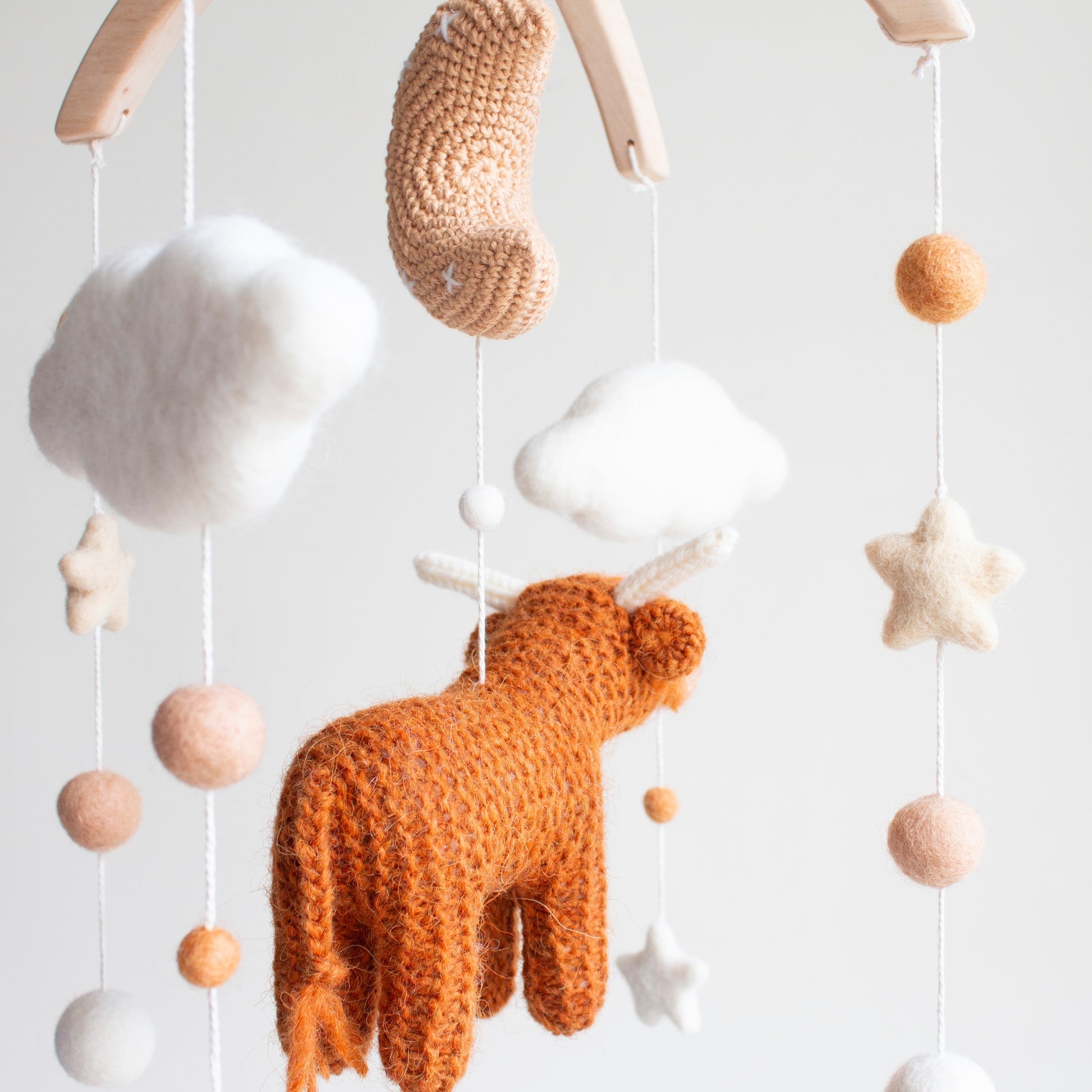 A rear-angle view of the artisan highland cow mobile highlighting the delicate balance of wool stars, felted balls, and a crochet moon in earthy, neutral tones.