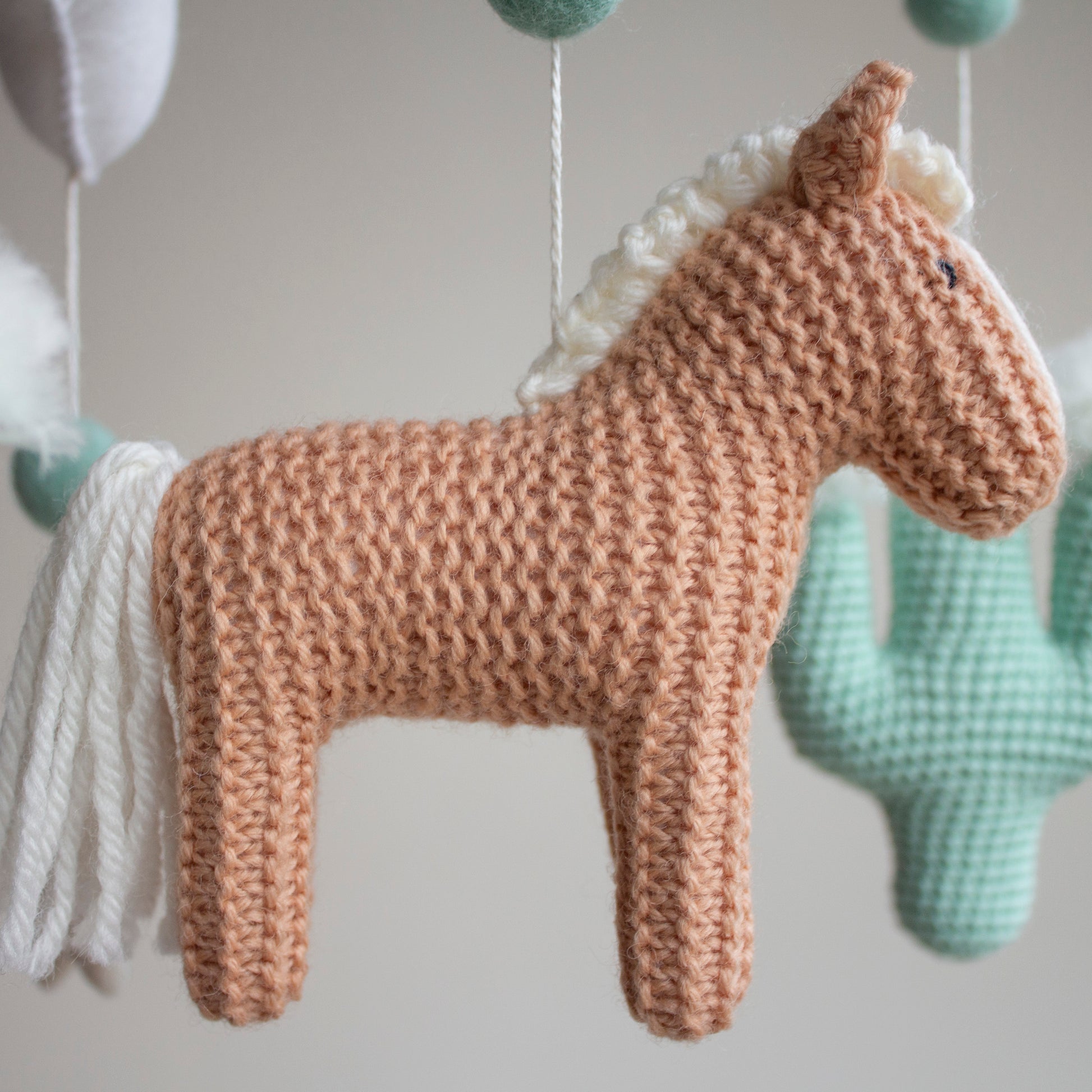 Close-up of a hand-knitted horse in a warm tan color with a soft white mane, illustrating the detailed artistry of our Western baby mobiles. This heirloom animal is designed to ignite wonder while maintaining a sophisticated, modern nursery aesthetic. By using non-toxic, certified materials, we provide a safe and high-quality decoration that is frequently listed on major UK baby registry platforms as a must-have item for new parents and stylish nursery rooms.