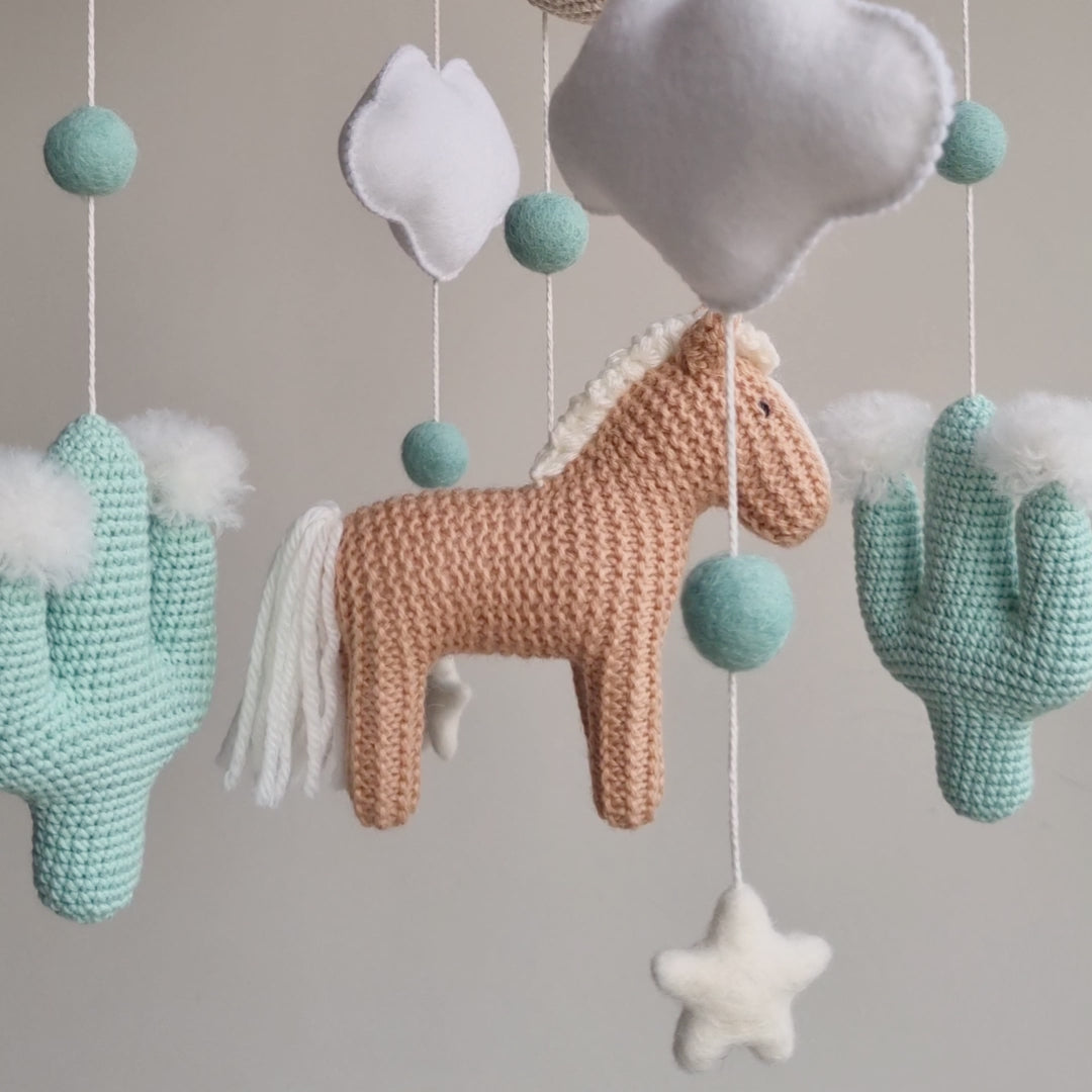 Artisan Western baby mobile featuring a tan hand-knitted horse, two mint green crochet cacti with white wool pom-poms, and a beige moon. Suspended from a sustainable wooden arch with ivory wool stars and clouds, this modern gender-neutral nursery decor is handcrafted in the UK from non-toxic materials. A prestigious heirloom-quality baby shower gift perfect for style-conscious mums-to-be looking for unique, ethical, and minimalist Scandi-inspired nursery accessories.