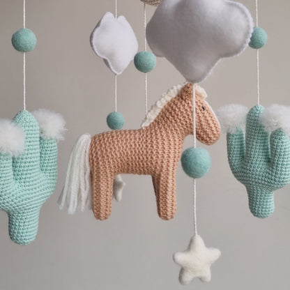 Artisan Western baby mobile featuring a tan hand-knitted horse, two mint green crochet cacti with white wool pom-poms, and a beige moon. Suspended from a sustainable wooden arch with ivory wool stars and clouds, this modern gender-neutral nursery decor is handcrafted in the UK from non-toxic materials. A prestigious heirloom-quality baby shower gift perfect for style-conscious mums-to-be looking for unique, ethical, and minimalist Scandi-inspired nursery accessories.